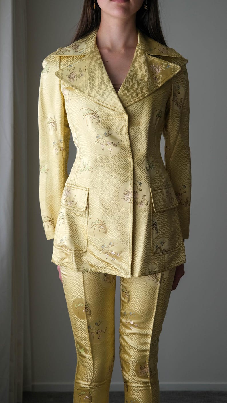 May include: A yellow suit with a floral and geometric pattern. The suit includes a double-breasted blazer with two front pockets and matching pants. The fabric has a subtle sheen, and the suit appears to be vintage.