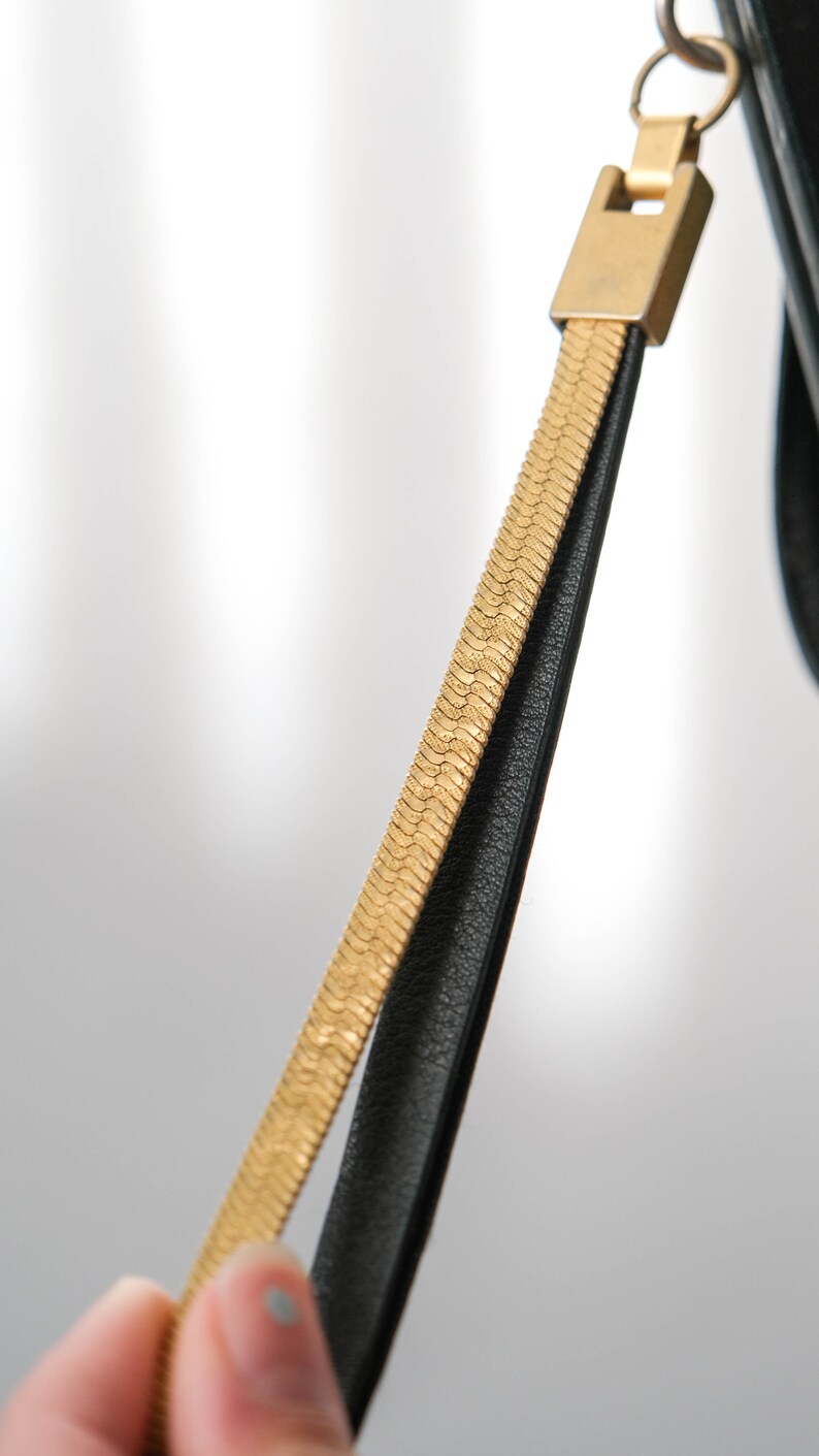 May include: A black leather strap with a gold chain detail. The strap is attached to a gold ring.