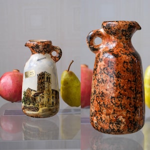 Op de afbeelding: Twee keramische kruiken met handvatten. Eén is wit met een kerkillustratie en het woord "ARGELIERS". De andere is oranje en zwart gespikkeld. Granaatappels en peren staan op de achtergrond.