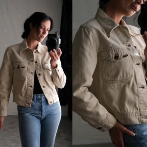 Puede incluir: Una chaqueta vaquera de color beige claro con cierre de botones y dos bolsillos delanteros. La chaqueta tiene un corte ligeramente recortado y está hecha de una tela suave y cómoda.