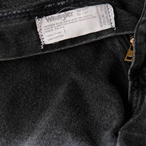 May include: A close-up of a black denim fabric with a white Wrangler label sewn into the fabric. The label includes the text "936WB", "Machine wash with like colors", "only non-chlorine bleach when needed", "tumble dry low", and "100% COTTON".
