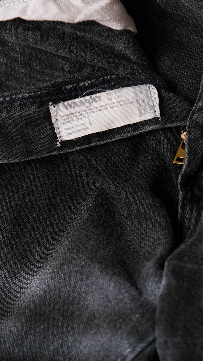 May include: A close-up of a black denim fabric with a white Wrangler label sewn into the fabric. The label includes the text "936WB", "Machine wash with like colors", "only non-chlorine bleach when needed", "tumble dry low", and "100% COTTON".