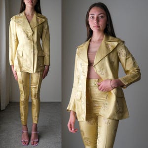 Puede incluir: Un traje amarillo de dos piezas con un estampado floral. El traje incluye una chaqueta con cuello y bolsillos y pantalones ajustados a juego. La tela tiene un brillo sutil y el atuendo se usa con sandalias rosas.