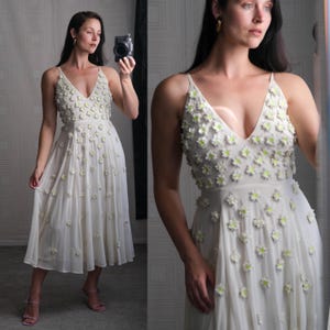 May include: A white midi dress with a V-neck and spaghetti straps, adorned with small, three-dimensional white flowers with yellow centers. The dress has a flowing skirt and is made of a lightweight fabric.
