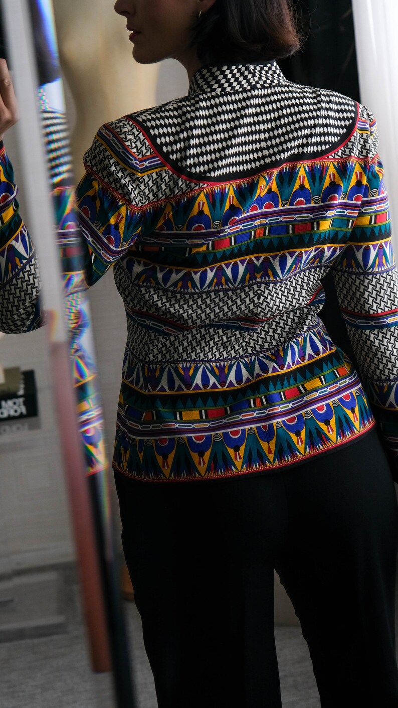 May include: A black and white patterned blouse with a colorful geometric design. The blouse has a high collar and long sleeves.