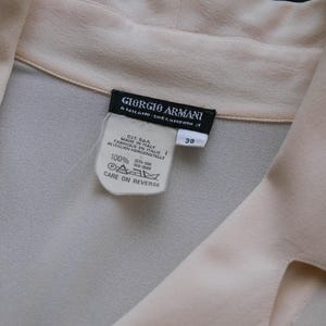 Peut inclure: Un chemisier en soie beige clair avec un col. L'&eacute;tiquette indique "GIORGIO ARMANI" et "MADE IN ITALY". Le chemisier porte une &eacute;tiquette de taille 38 et des instructions d'entretien.