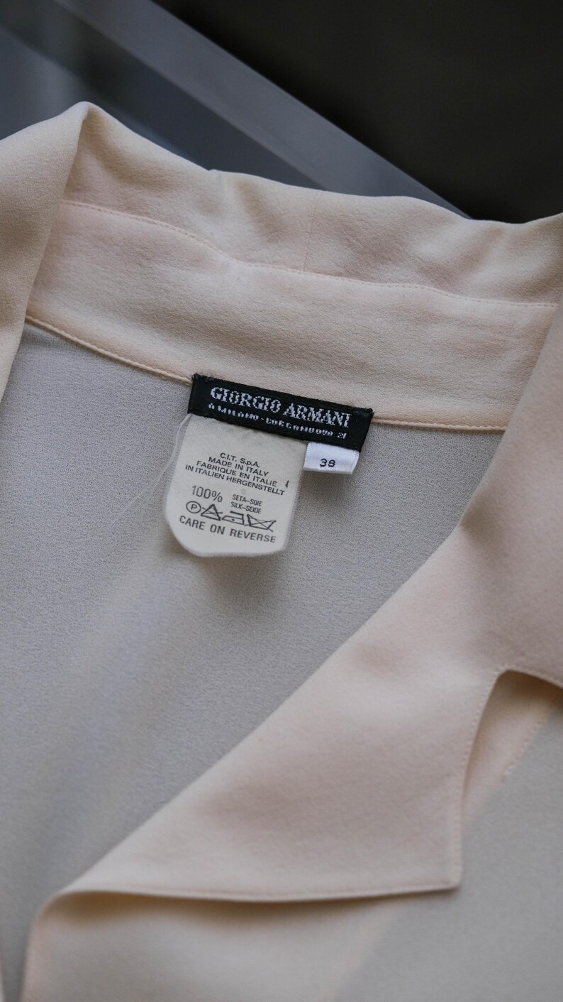 Peut inclure: Un chemisier en soie beige clair avec un col. L'&eacute;tiquette indique "GIORGIO ARMANI" et "MADE IN ITALY". Le chemisier porte une &eacute;tiquette de taille 38 et des instructions d'entretien.