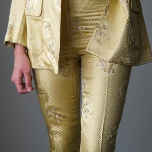 May include: A two-piece yellow suit with a matching jacket and pants. The jacket has two front pockets and the pants have a geometric pattern and floral embroidery. The suit is made of a shiny fabric.