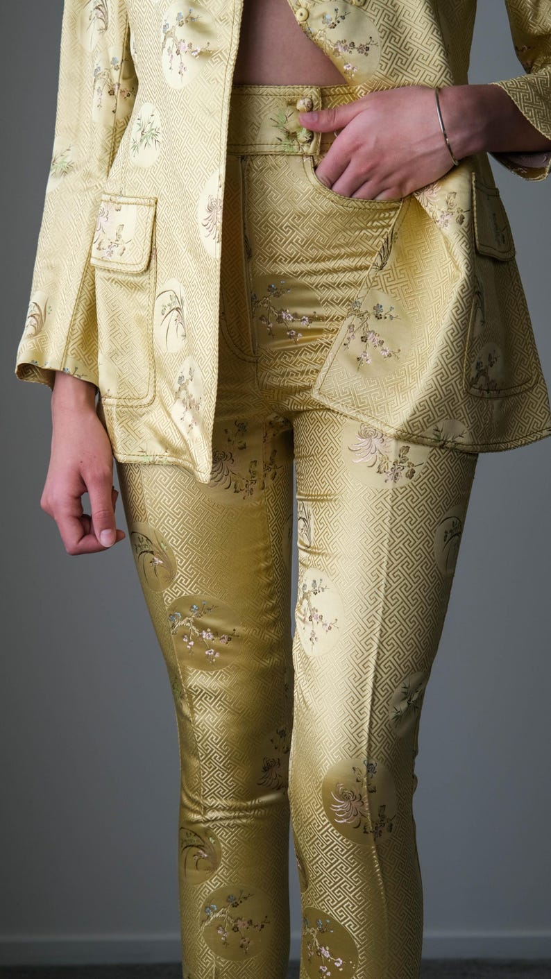 May include: A two-piece yellow suit with a matching jacket and pants. The jacket has two front pockets and the pants have a geometric pattern and floral embroidery. The suit is made of a shiny fabric.