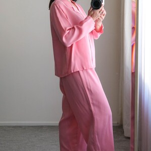 May include: A light pink pajama set, including a long-sleeved button-down shirt and wide-leg pants. The outfit is paired with light pink heeled mules. The person is holding a black camera.