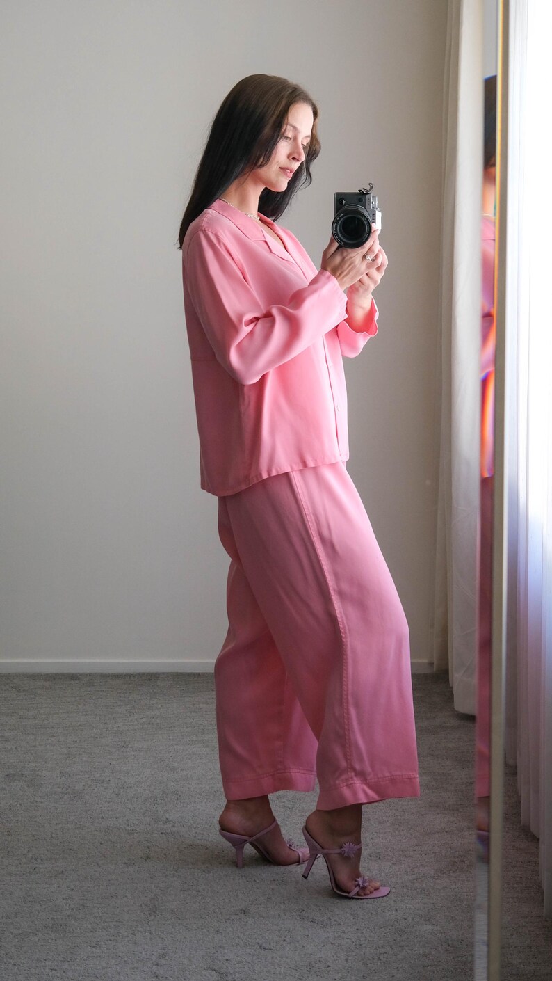 May include: A light pink pajama set, including a long-sleeved button-down shirt and wide-leg pants. The outfit is paired with light pink heeled mules. The person is holding a black camera.