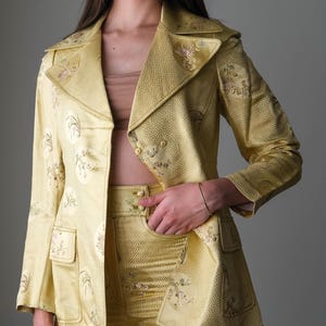 May include: A golden yellow suit with a floral pattern. The suit includes a blazer with a notched collar and two pockets, and matching high-waisted pants. The fabric has a subtle sheen. The suit is a vintage style.