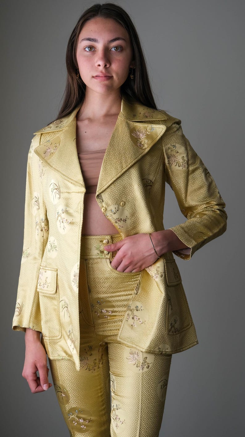 May include: A golden yellow suit with a floral pattern. The suit includes a blazer with a notched collar and two pockets, and matching high-waisted pants. The fabric has a subtle sheen. The suit is a vintage style.