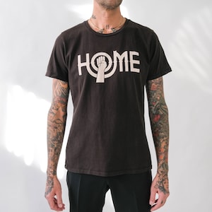 Może przedstawiać: Czarny T-shirt z białym nadrukiem pięści w kółku z napisem "HOME" wokół.