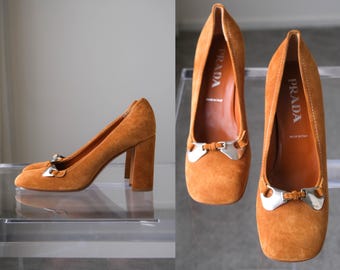 Puede incluir: Un par de zapatos de tacón de bloque en ante color canela con punta cuadrada y un detalle de hebilla de metal plateado en el empeine. La plantilla es de cuero canela con el logo de Prada y el texto Made in Italy impresos en blanco.