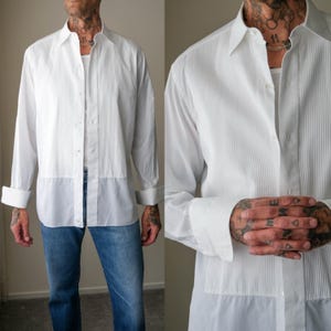 May include: A white long-sleeved button-down shirt featuring a vertical pleated bib front design, a classic collar, and cuffed sleeves. The shirt is worn over a white undershirt and paired with blue denim jeans, showcasing a crisp, structured look.