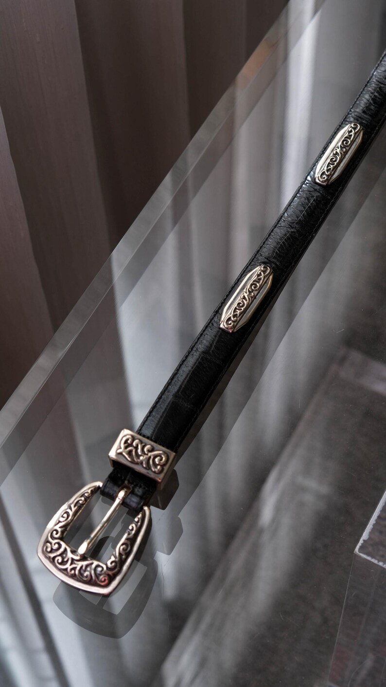 May include: Black leather belt with silver-tone metal buckle and decorative accents. The buckle and accents have an ornate, swirling design. The belt is displayed on a reflective surface.