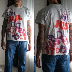 May include: White t-shirt featuring a graphic print of the word "JUST" in red, purple and white. The print is layered and has a spray paint effect. The shirt is being worn by a person with tattoos on their arms.