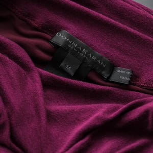 May include: Close-up of a deep magenta-colored garment with soft folds. A black label with the text "DONNA KARAN NEW YORK" and the size "M" is visible. The label also indicates the item was made in Italy.