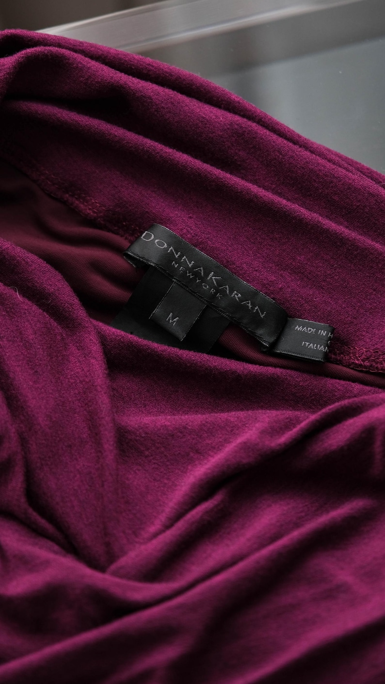 May include: Close-up of a deep magenta-colored garment with soft folds. A black label with the text "DONNA KARAN NEW YORK" and the size "M" is visible. The label also indicates the item was made in Italy.