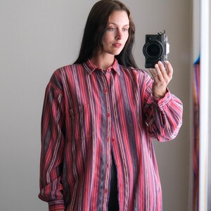May include: A button-down shirt with vertical stripes in shades of red, purple, and beige. The shirt has a chest pocket and long sleeves. The model is wearing the shirt with dark pants.