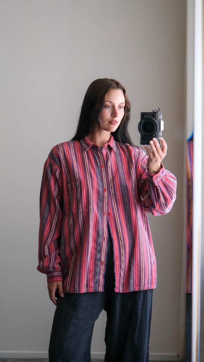 May include: A button-down shirt with vertical stripes in shades of red, purple, and beige. The shirt has a chest pocket and long sleeves. The model is wearing the shirt with dark pants.