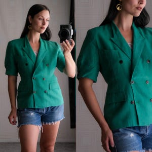 May include: A double-breasted, emerald green blazer with short sleeves. The blazer features two rows of dark buttons, a notched lapel, and two front pockets. The model is wearing denim shorts. The blazer is made of a textured fabric.