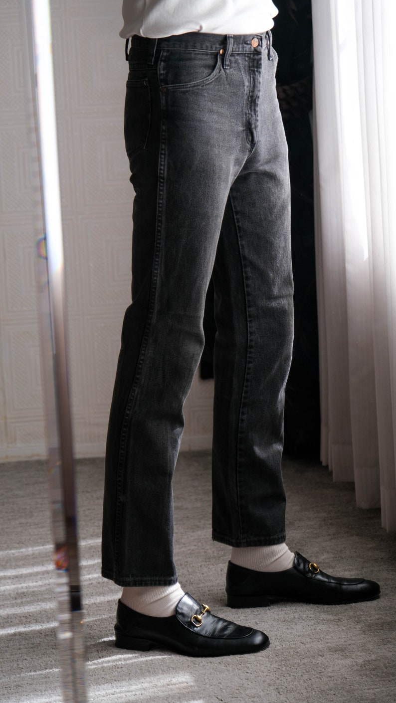 May include: A person wearing black jeans and black loafers with gold accents. The person is standing on a carpeted floor.