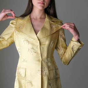 May include: A yellow suit with a floral pattern. The suit includes a collared blazer with two front pockets and matching pants. The fabric has a subtle geometric design. The outfit is a vintage style.