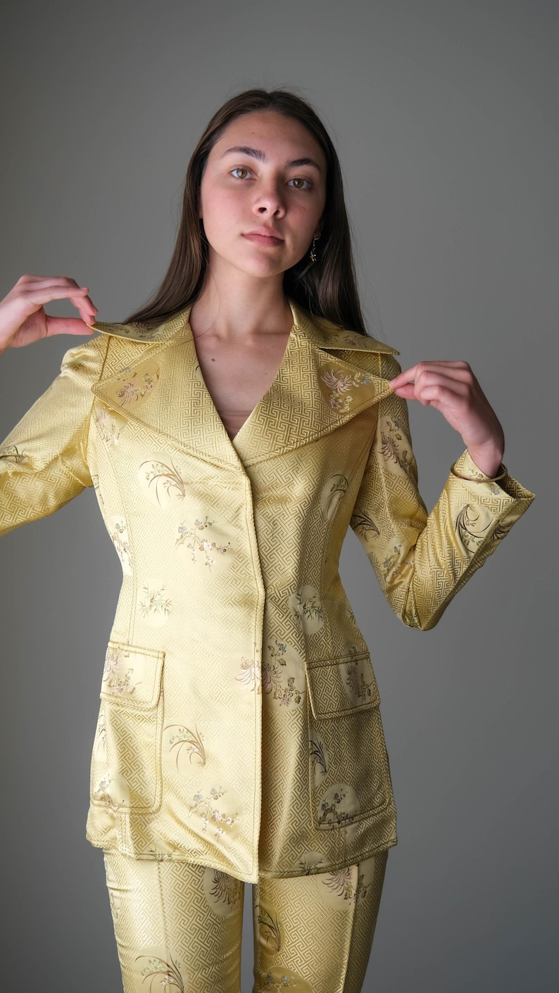 May include: A yellow suit with a floral pattern. The suit includes a collared blazer with two front pockets and matching pants. The fabric has a subtle geometric design. The outfit is a vintage style.