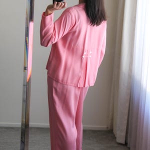 May include: A light pink two-piece outfit, consisting of a long-sleeved top and wide-leg pants. The top has a pleated back detail with small white buttons. The outfit is paired with pink heeled sandals. The image is taken in front of a mirror.
