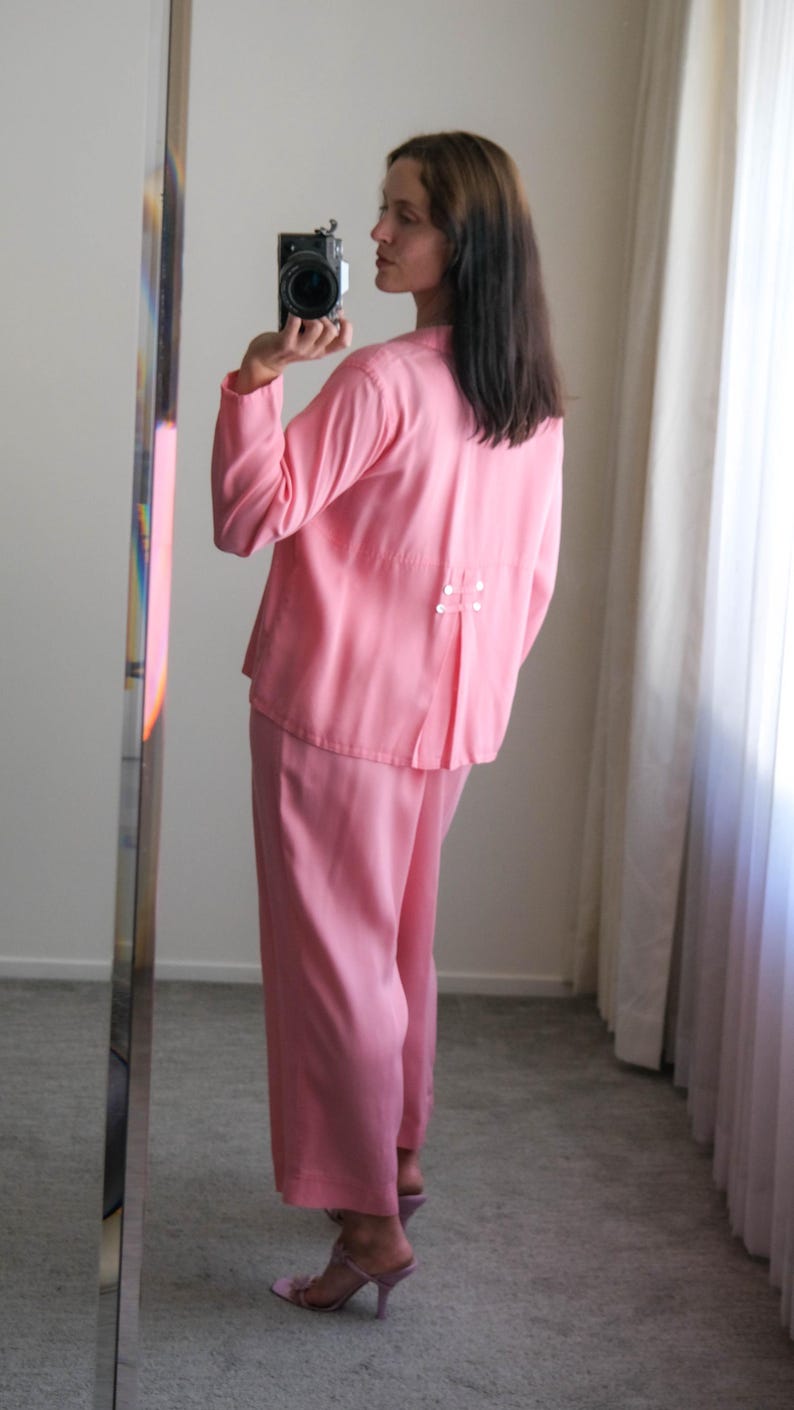 May include: A light pink two-piece outfit, consisting of a long-sleeved top and wide-leg pants. The top has a pleated back detail with small white buttons. The outfit is paired with pink heeled sandals. The image is taken in front of a mirror.