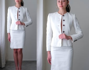 May include: A white skirt suit featuring a tailored jacket with dark buttons and a matching knee-length skirt. The jacket has a round collar and small pockets. The suit is made of a textured fabric, creating a professional and polished look.