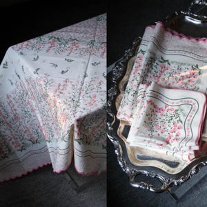May include: A white tablecloth and matching napkins with a floral pattern in pink and green. The tablecloth has a scalloped edge with pink trim. The napkins are neatly stacked on a silver tray.