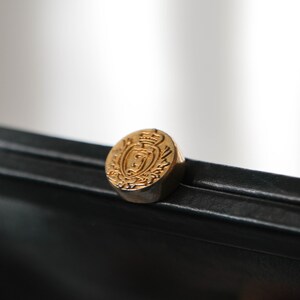 May include: A black leather clutch with a gold-toned clasp featuring a crest design.