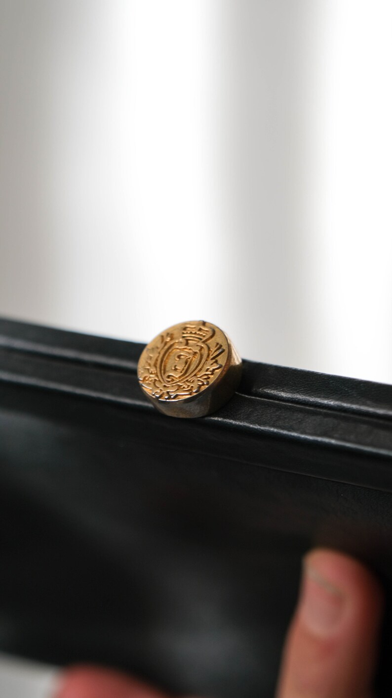 May include: A black leather clutch with a gold-toned clasp featuring a crest design.