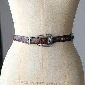 K&ouml;nnte beinhalten: Ein brauner Lederg&uuml;rtel mit silberner Schnalle und dekorativen Akzenten. Der G&uuml;rtel wird auf einer Schaufensterpuppe pr&auml;sentiert. Die Schnalle hat eine rechteckige Form mit einem rautenf&ouml;rmigen Goldakzent. Der G&uuml;rtel ist etwa 2,5 cm breit.