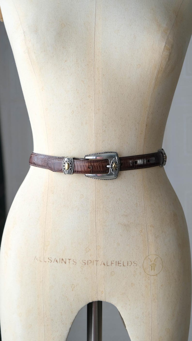 K&ouml;nnte beinhalten: Ein brauner Lederg&uuml;rtel mit silberner Schnalle und dekorativen Akzenten. Der G&uuml;rtel wird auf einer Schaufensterpuppe pr&auml;sentiert. Die Schnalle hat eine rechteckige Form mit einem rautenf&ouml;rmigen Goldakzent. Der G&uuml;rtel ist etwa 2,5 cm breit.