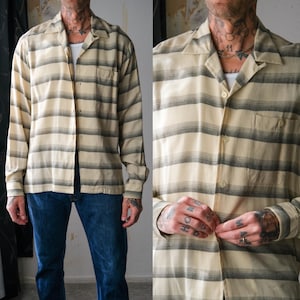 Puede incluir: Camisa de manga larga con cuello de campamento. La camisa presenta rayas horizontales en tonos beige y gris. Tiene un bolsillo en el pecho y se lleva abierta sobre una camiseta interior blanca. El modelo lleva vaqueros azules.