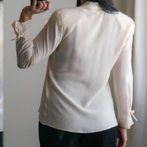 Peut inclure: Un chemisier cr&egrave;me &agrave; manches longues et col montant. Les manches sont fronc&eacute;es aux poignets et nou&eacute;es avec de petits n&oelig;uds. Le chemisier est associ&eacute; &agrave; un pantalon noir. Un appareil photo est tenu dans la main.