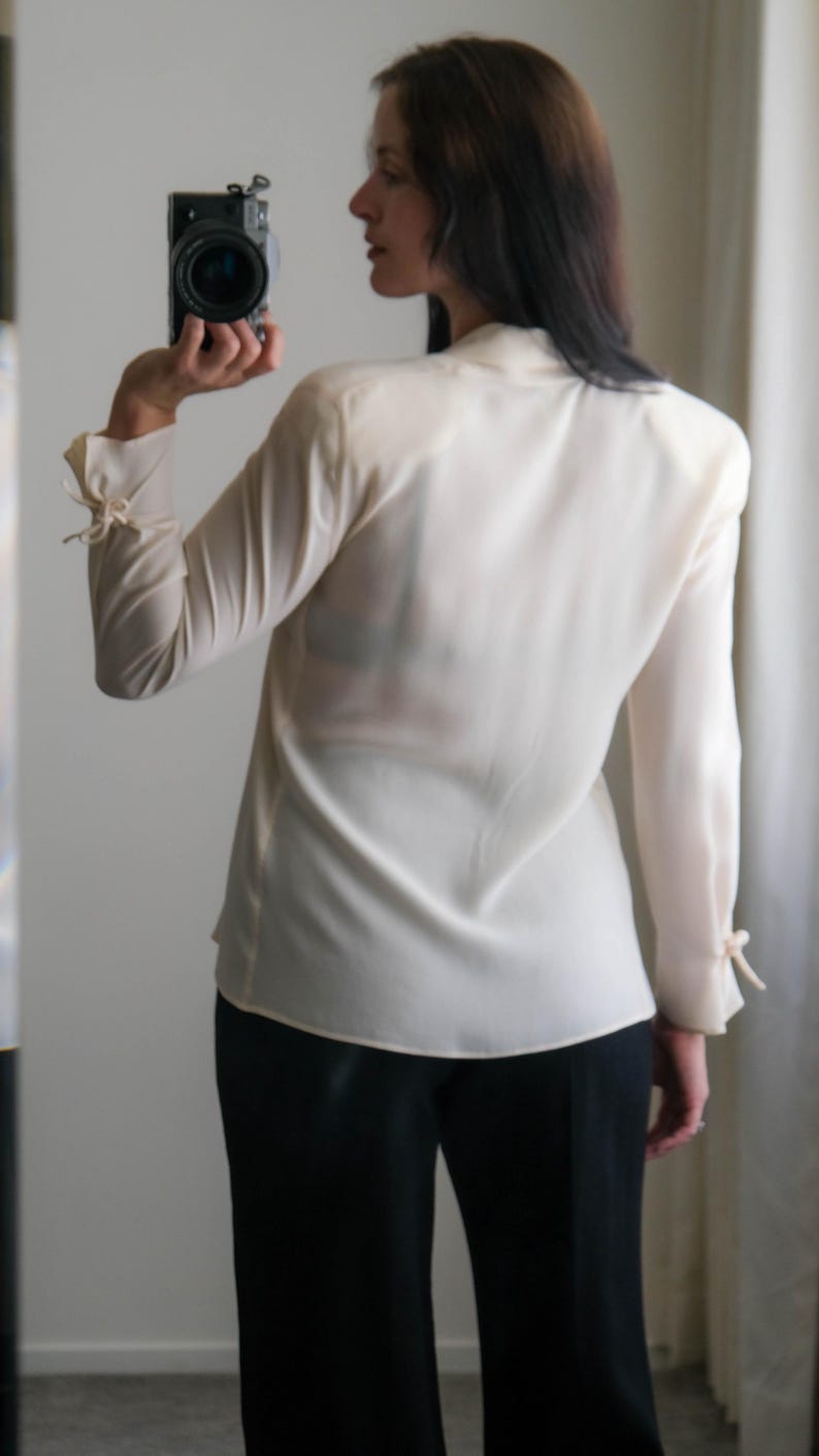 Peut inclure: Un chemisier cr&egrave;me &agrave; manches longues et col montant. Les manches sont fronc&eacute;es aux poignets et nou&eacute;es avec de petits n&oelig;uds. Le chemisier est associ&eacute; &agrave; un pantalon noir. Un appareil photo est tenu dans la main.