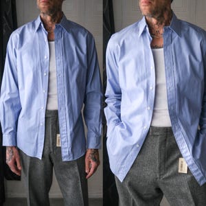 Op de afbeelding: Een lichtblauw overhemd met lange mouwen, gecombineerd met een grijze broek. Het overhemd is opengeknoopt en onthult een wit ondershirt. Het overhemd heeft een klassieke kraag en manchetten met knopen. De outfit wordt gecompleteerd door een grijze broek.