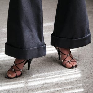 May include: Black high-heeled sandals with rhinestone accents and black flared pants with a cuffed hem. The shoes have thin straps and a stiletto heel. The pants are a dark color.