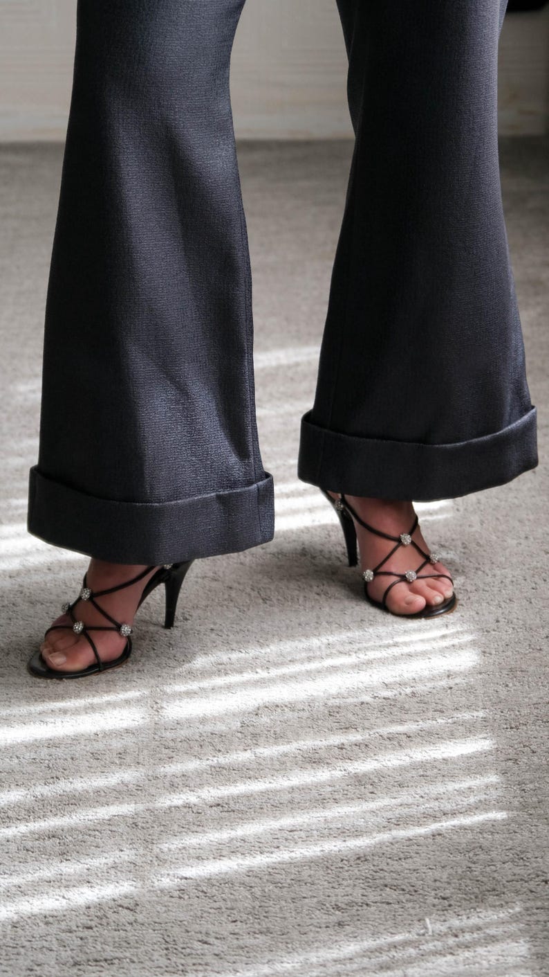 May include: Black high-heeled sandals with rhinestone accents and black flared pants with a cuffed hem. The shoes have thin straps and a stiletto heel. The pants are a dark color.