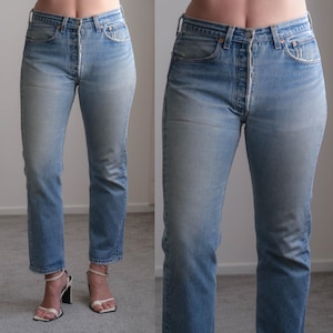 May include: High-waisted light blue denim jeans featuring a classic five-pocket design, a button-fly closure, and a straight-leg cut. The denim has a faded wash with whiskering details at the hips and a straight hemline at the ankles.
