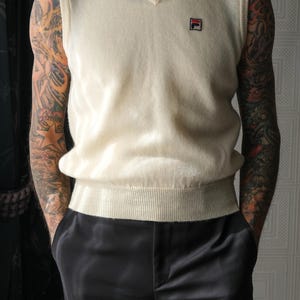 Peut inclure: Un gilet de pull cr&egrave;me &agrave; col en V avec un petit logo sur la poitrine. Le gilet est associ&eacute; &agrave; un pantalon en satin noir. La tenue est de style classique et minimaliste.