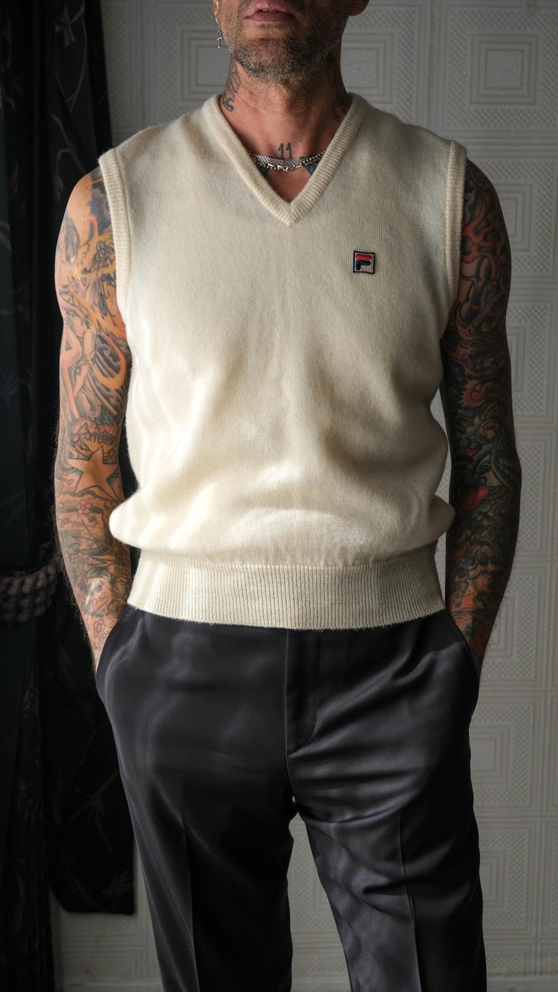 Peut inclure: Un gilet de pull cr&egrave;me &agrave; col en V avec un petit logo sur la poitrine. Le gilet est associ&eacute; &agrave; un pantalon en satin noir. La tenue est de style classique et minimaliste.