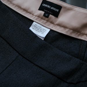 May include: Close-up of a dark gray fabric garment with a beige lining. The label reads "GIORGIO ARMANI MADE IN ITALY" with the size "48". A smaller tag provides the RN number and address for Giorgio Armani Corp. in New York.
