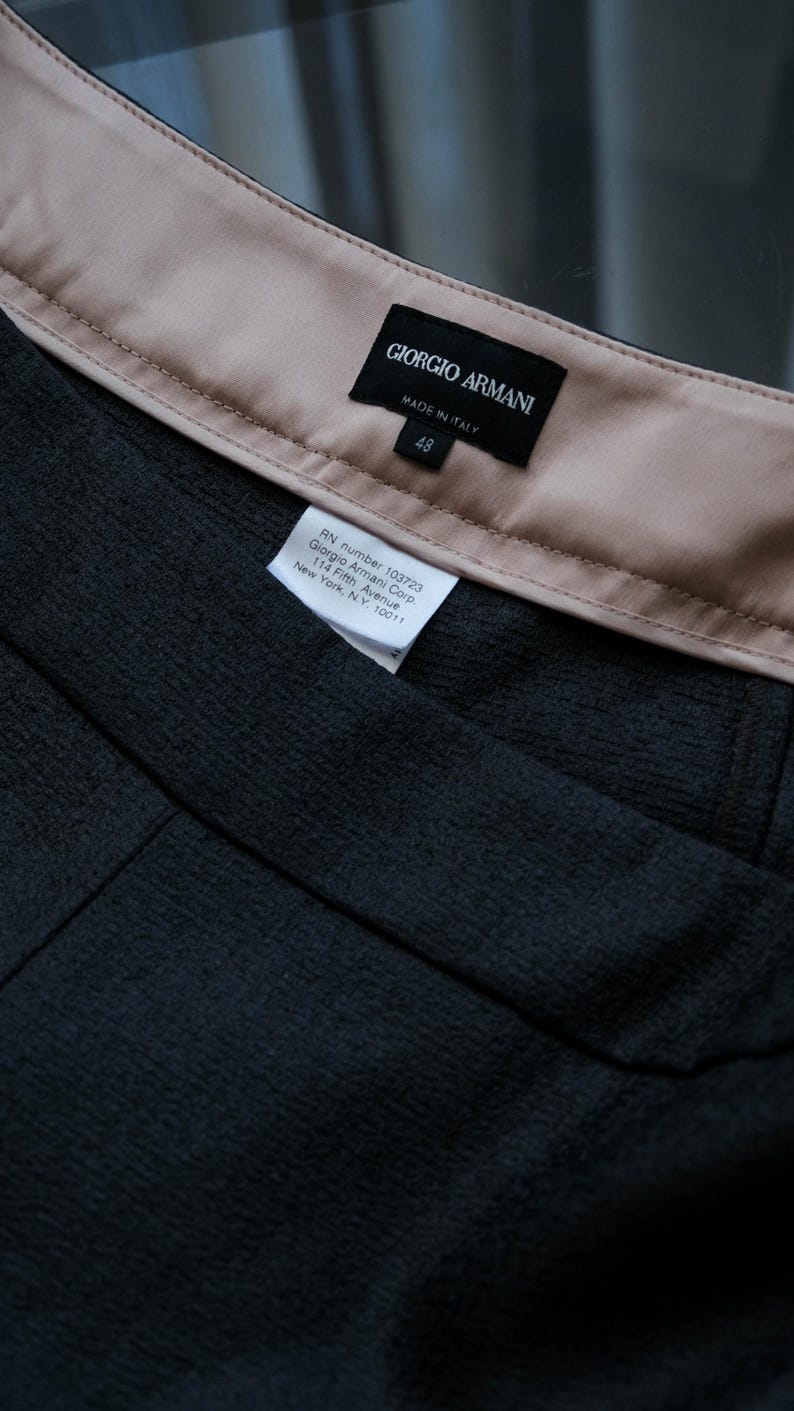 May include: Close-up of a dark gray fabric garment with a beige lining. The label reads "GIORGIO ARMANI MADE IN ITALY" with the size "48". A smaller tag provides the RN number and address for Giorgio Armani Corp. in New York.