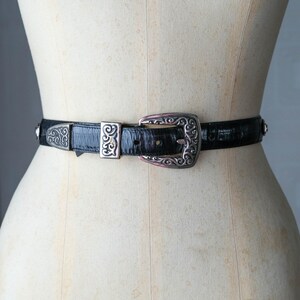 May include: Black belt with a silver buckle and accents. The belt is made of a textured material and features a decorative buckle with an ornate design. The belt is displayed on a mannequin.